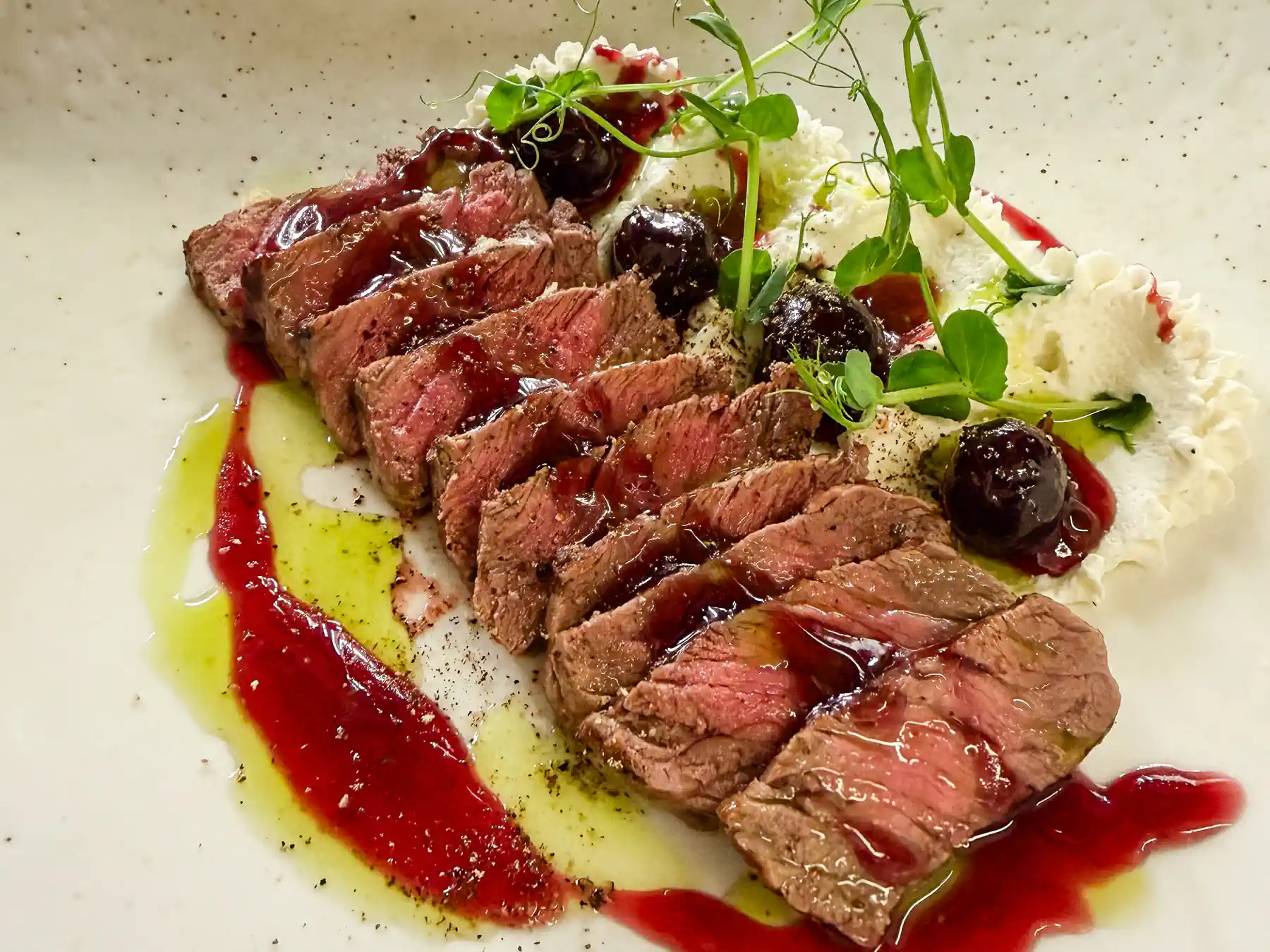 Auf einem hellen Teller liegen gleichmässig geschnittene Scheiben rosa gebratenen Rindfleischs in einer Reihe angerichtet. Glänzende dunkle Tupfer und rote Saucenspuren ziehen sich über den Teller, begleitet von weissen, luftigen Cremenocken. Frische grüne Sprossen setzen leichte Akzente und geben dem Gericht eine elegante, mediterrane Anmutung.