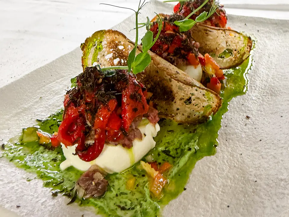 Auf einer länglichen, hellen Platte liegt ein leuchtend grünes Kräuterbett, darauf mehrere halbmondförmig geöffnete Mozzarella-Säckchen. In der Mitte quillt eine dunkle, fein gewürfelte Füllung hervor, gekrönt von glänzenden, tiefroten Paprikastreifen. Kleine Würfel und Tropfen von Sauce setzen zusätzliche Akzente, während frische Sprossen dem Arrangement Höhe und Leichtigkeit verleihen.