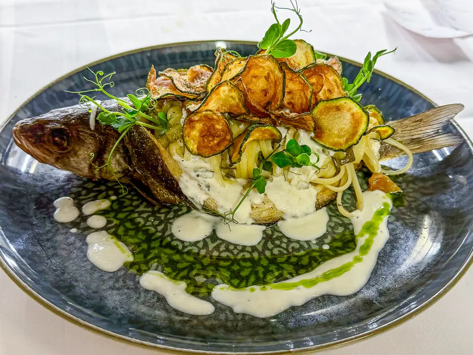 Auf einem großen, dunkel schimmernden Teller liegt ein ganzer, goldbraun gebratener Fisch seitlich angerichtet. Daneben und darunter schmiegen sich helle Linguine in cremiger Sauce, teilweise bedeckt von knusprig frittierten Zucchinischeiben, die sich wie ein lockerer Turm darüberlegen. Helle Saucenspiegel und ein leuchtend grünes Kräuteröl ziehen sich dekorativ über den Teller, während frische Sprossen dem Gericht eine elegante, mediterrane Leichtigkeit verleihen.