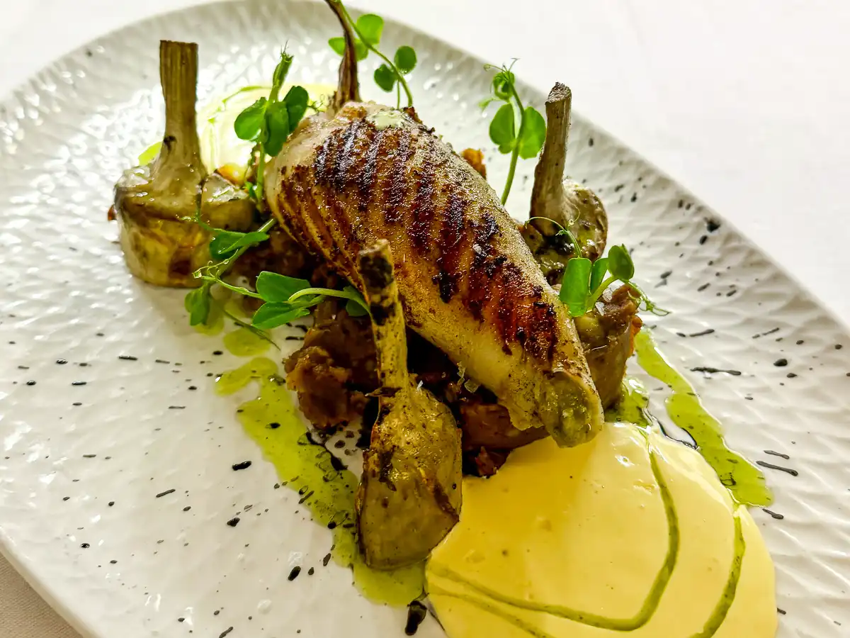 Auf einem hellen, strukturierten Teller liegt ein ganzes, goldbraun gegrilltes Kalmarstück mittig angerichtet. Es ruht auf einem Bett aus cremigem Püree, umgeben von kleinen Artischocken und glänzenden Saucenspiegeln. Frische Kräuter und grüne Öltropfen setzen lebendige Akzente und verleihen dem Gericht eine elegante, mediterrane Optik.