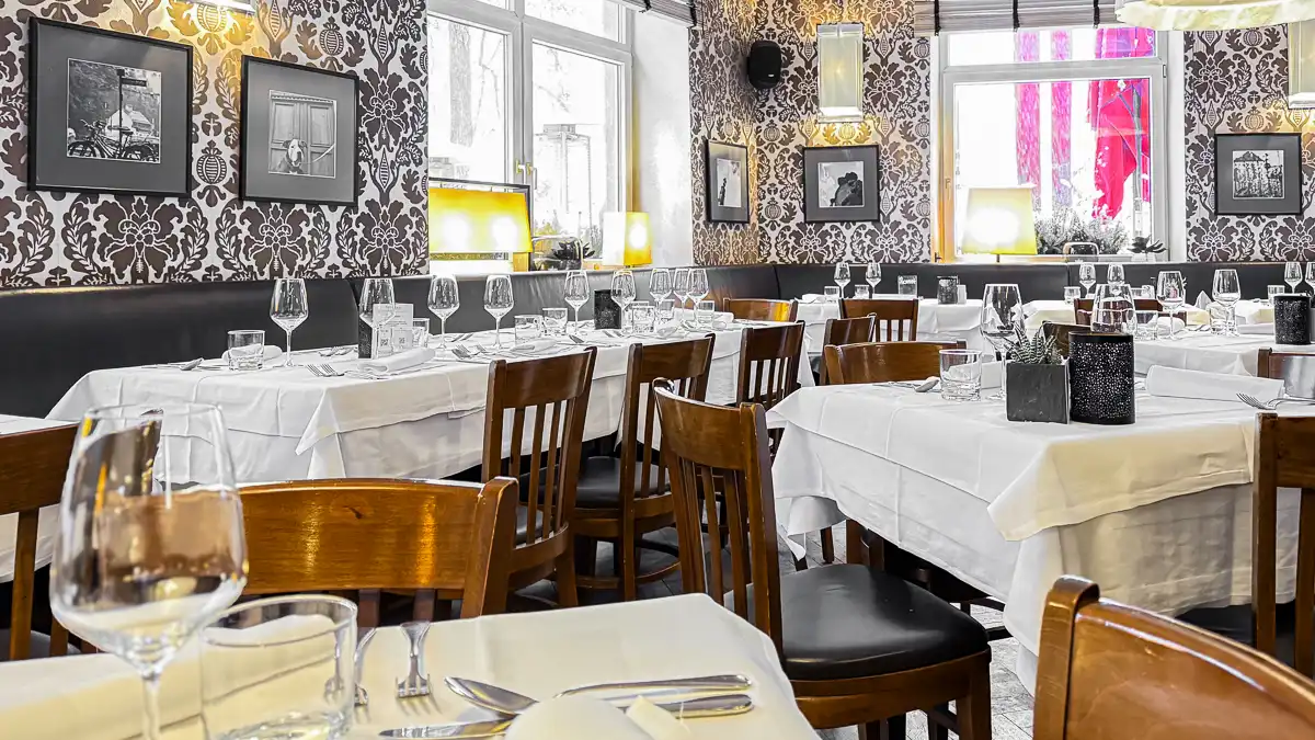 Ein stilvoll eingerichteter Gastraum mit weiß gedeckten Tischen, eleganten Weingläsern und warmem, gedämpftem Licht. Dunkle Holzstühle und gemusterte Tapeten verleihen dem Raum eine klassische, gemütliche Atmosphäre, während große Fenster für Helligkeit und Offenheit sorgen.