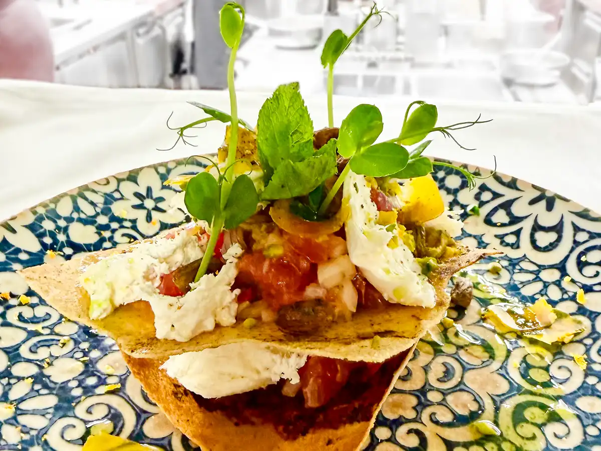 Auf einem dekorativ gemusterten Teller ist ein knusprig geschichtetes Arrangement aus dünnen Brotplatten aufgebaut. Dazwischen und darauf liegen cremige, helle Elemente und fein gewürfelte Zutaten in rötlichen Tönen. Frische grüne Sprossen ragen nach oben und verleihen dem Gericht Höhe, während kleine gelbe und grüne Akzente das Gesamtbild lebendig und mediterran wirken lassen.