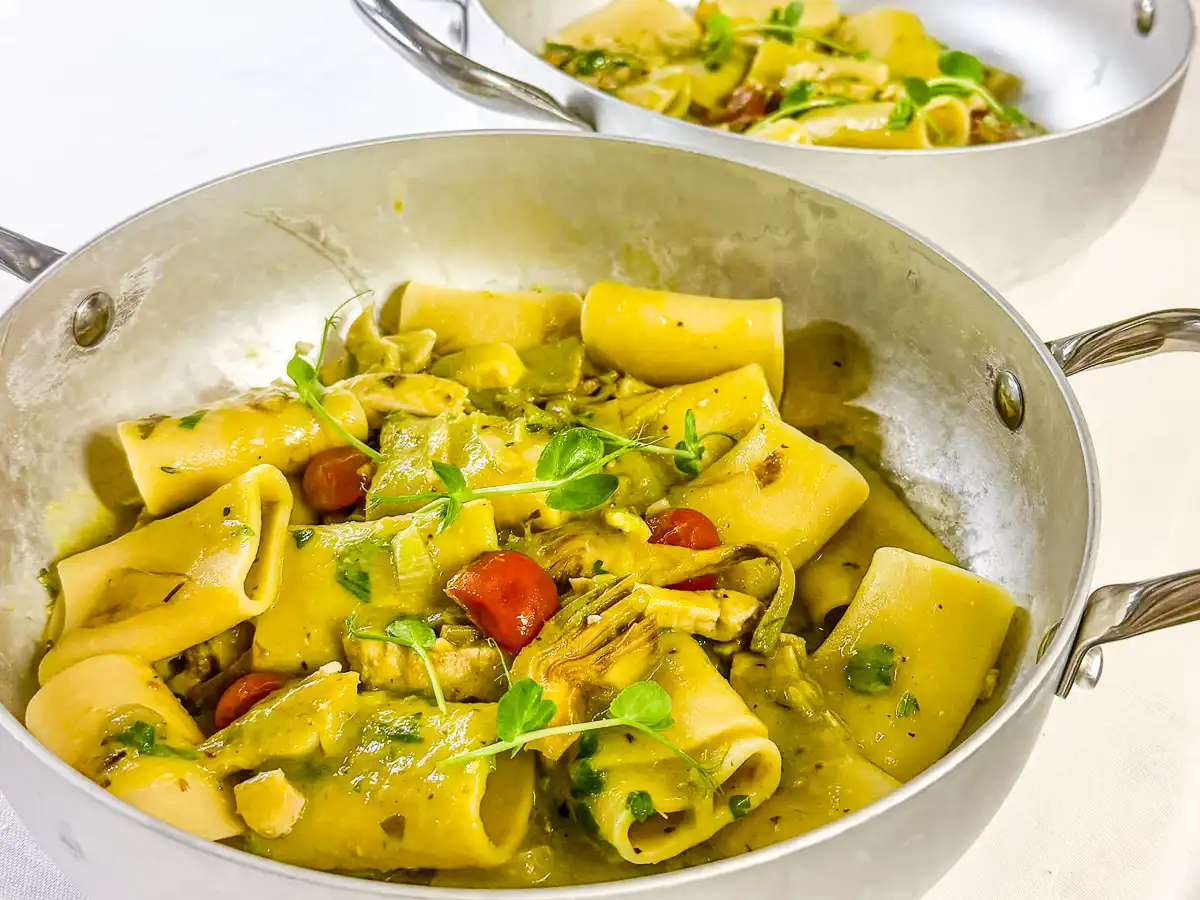In einer silbernen Pfanne sind große, röhrenförmige Paccheri in einer cremigen, gelblich-grünen Sauce angerichtet. Dazwischen setzen rote Tomaten, grüne Gemüsestücke und knusprige Elemente farbige Akzente, während frische Kräuter dem Gericht eine lebendige, mediterrane Frische verleihen.