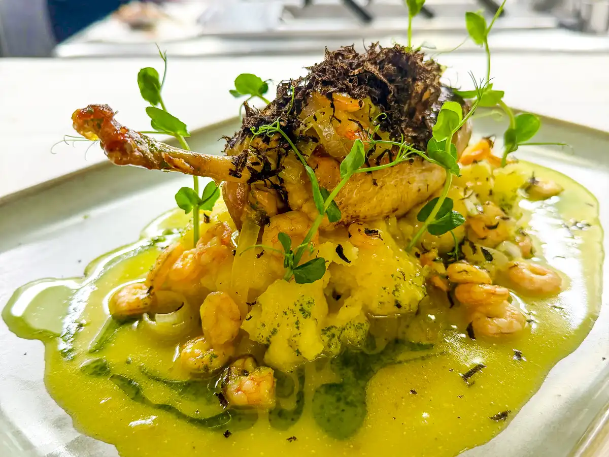 Auf einem hellen Teller ist eine knusprig gebratene Perlhuhnbrust auf einem Bett aus cremigem Kartoffelpüree angerichtet. Darum verteilt liegt eine leuchtend gelbgrüne Sauce mit kleinen Garnelen, darüber feine schwarze Trüffelspäne und frische Sprossen, die dem Gericht Höhe und Frische verleihen.