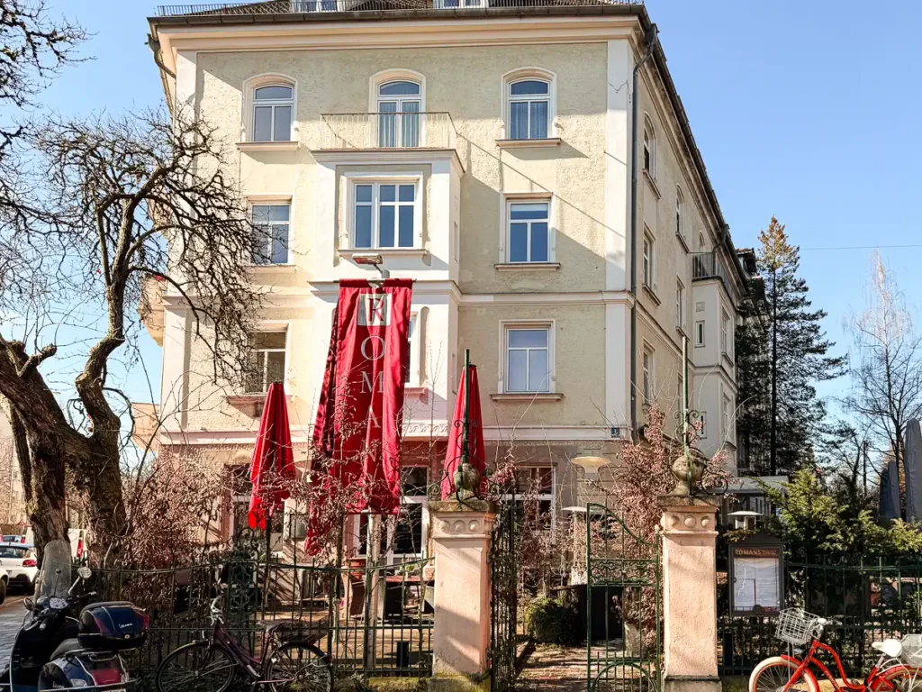 Ein elegantes, helles Gebäude mit klassischer Fassade, davor eine kleine, eingefasste Terrasse mit geschlossenen roten Sonnenschirmen. Ein schmiedeeisernes Tor führt in den Außenbereich, während Bäume und Fahrräder dem Eingangsbereich eine lebendige, urbane Note verleihen.