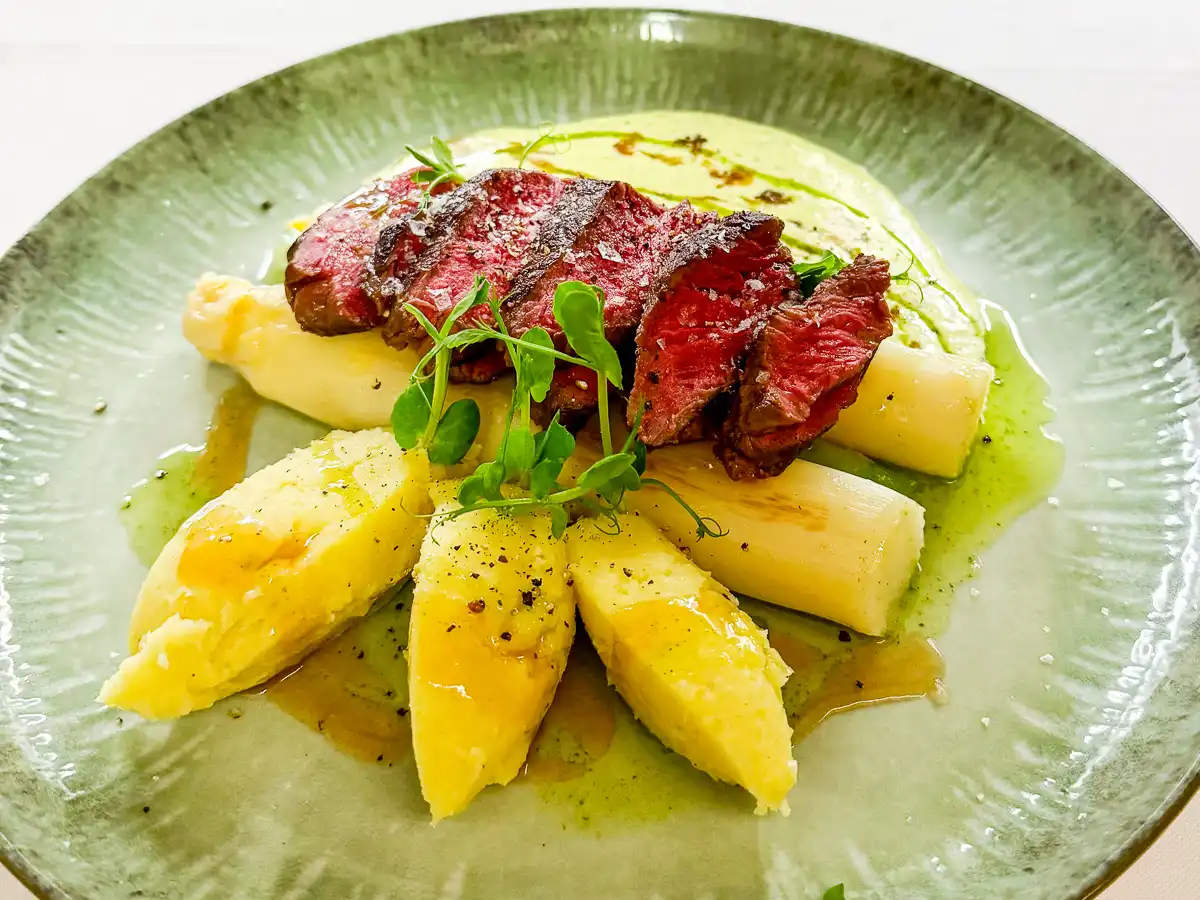 Auf einem großen, grünen Teller sind rosa aufgeschnittene Fleischscheiben fächerartig angerichtet. Darunter und daneben liegen helle Spargelstücke und goldgelbe Kartoffelkomponenten, umgeben von glänzenden Saucen und grünen Öltropfen. Frische Kräuter setzen leichte, elegante Akzente und verleihen dem Gericht eine klare, mediterrane Ausstrahlung.