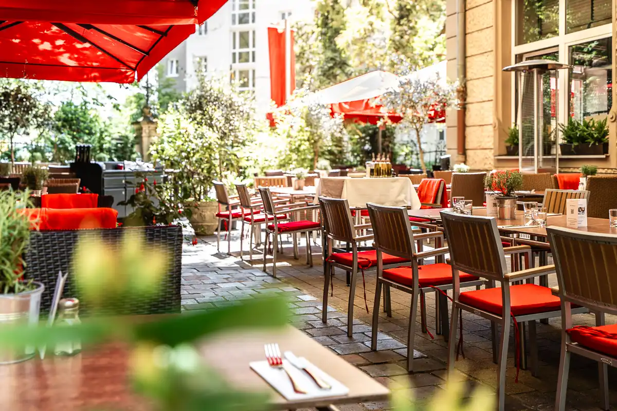 Eine sonnige Außenterrasse mit stilvoll gedeckten Tischen und Stühlen mit roten Polstern. Große rote Sonnenschirme spenden Schatten, während Pflanzen und helle Fassaden eine einladende, mediterrane Atmosphäre schaffen.