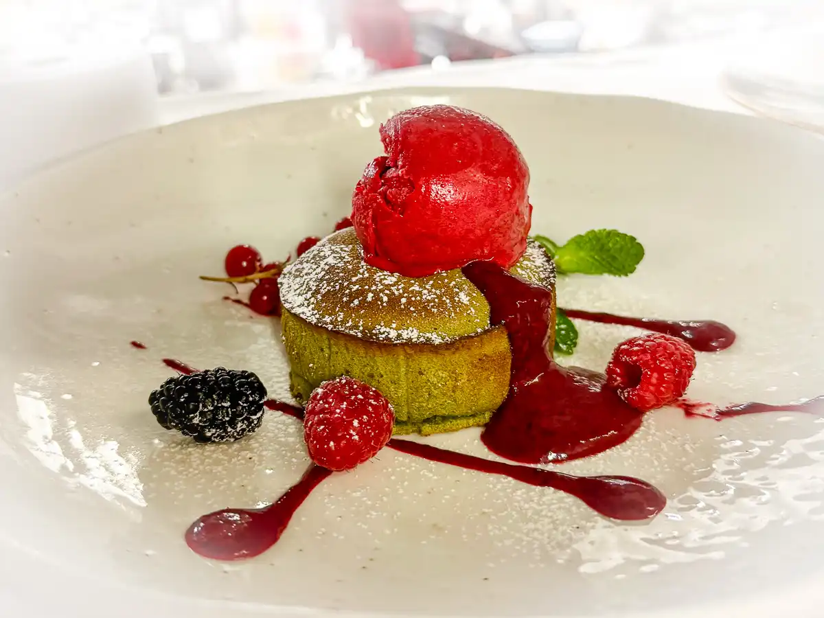 Auf einem großen, hellen Teller steht ein runder, grüner Küchlein mit leicht aufgerissener Oberfläche, darauf eine Kugel leuchtend rotes Sorbet. Rote Beerensauce fließt über das Dessert und verteilt sich dekorativ auf dem Teller, begleitet von frischen Beeren und einem Hauch Puderzucker.