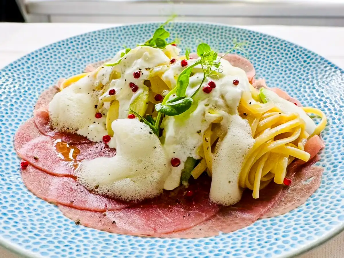 Auf einem hellblau gemusterten Teller liegt ein Nest aus hellgelben Tagliolini, umgeben von dünnen, rosafarbenen Carpaccio-Scheiben. Darüber fließt eine luftige, helle Schaumsauce, akzentuiert von grünen Spargelstücken, kleinen roten Pfefferkörnern und frischen Sprossen.