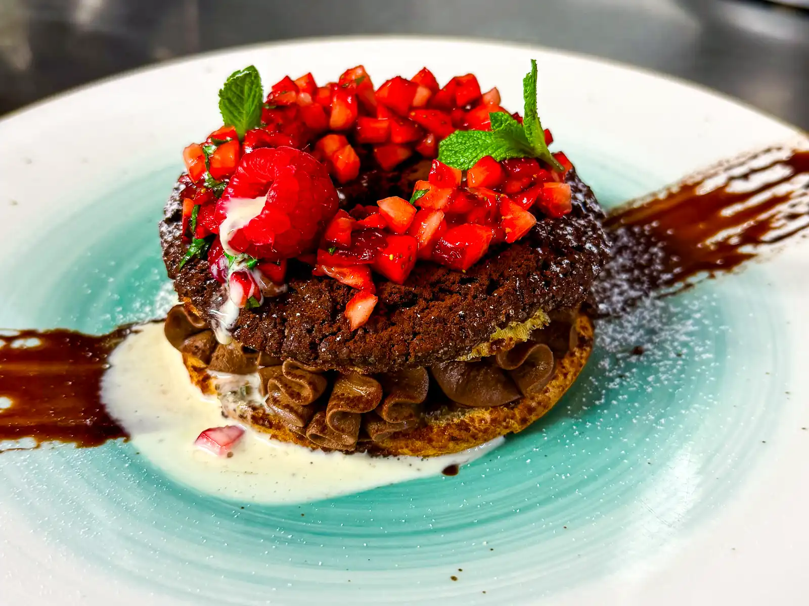 Auf einem großen, hellen Teller liegt ein rundes, schokoladiges Dessert mit knuspriger Struktur. Darauf häufen sich fein gewürfelte, leuchtend rote Erdbeeren, akzentuiert von frischer Minze. Eine helle Creme und dunkle Schokoladensauce umrahmen das Dessert und verleihen ihm eine elegante, kontrastreiche Optik.