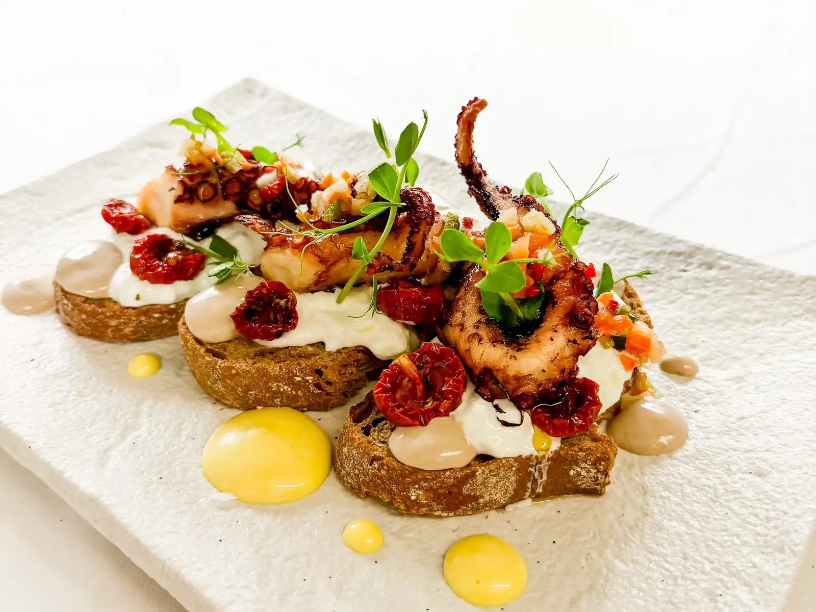 Auf einer hellen, rechteckigen Platte liegen geröstete Brotscheiben, darauf angerichtet zarte Stücke von gegrilltem Oktopus. Cremige, helle Tupfer und glänzende rote Akzente verteilen sich darüber, während frische Kräuter und kleine Gemüsestücke dem Gericht Lebendigkeit und mediterrane Frische verleihen.