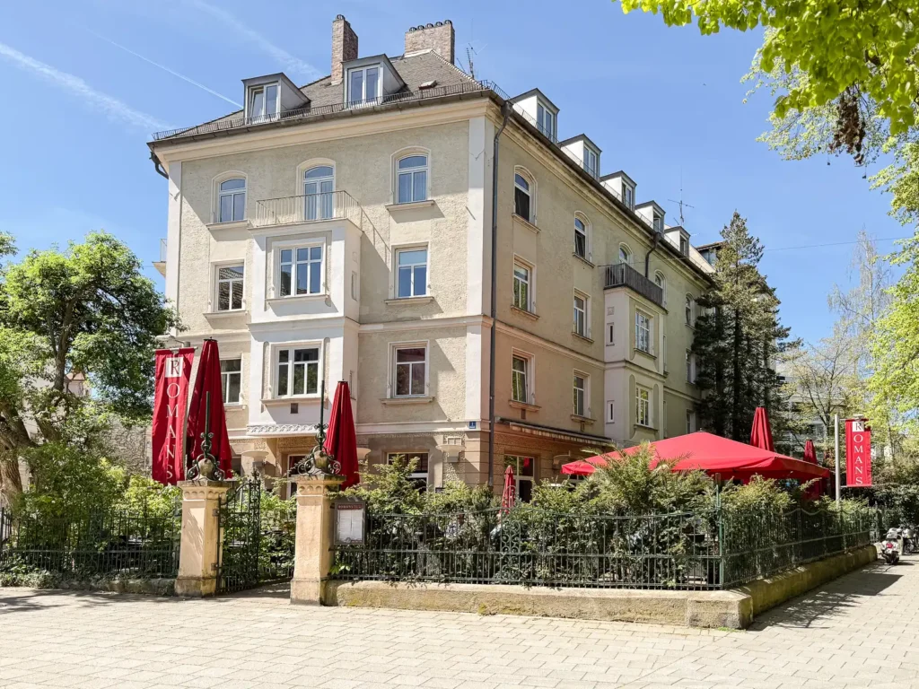 Ristorante ROMANS, eine helle Villa mit Erkern und Dachgauben, umgeben von einem Gartenzaun, Bäumen und roten Sonnenschirmen auf einer Terrasse in München Neuhausen-Nymphenburg.