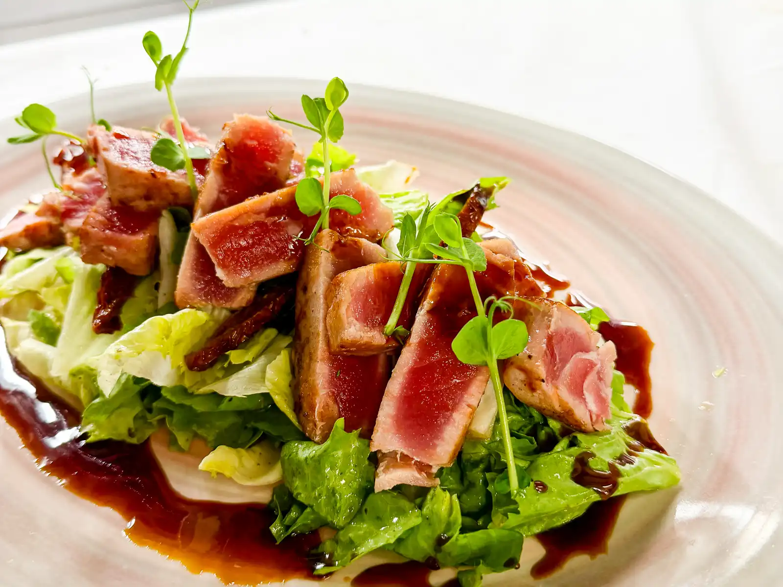 Auf einem hellen Teller sind dick geschnittene, rosa Thunfischscheiben locker auf einem Bett aus grünen Blattsalaten angerichtet. Eine dunkle, glänzende Sauce umrahmt das Gericht, während frische Sprossen und feine Kräuter leichte, elegante Akzente setzen.