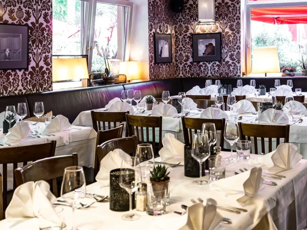 Festlich gedeckte Tische in einem Restaurant mit weissen Tischdecken, gefalteten Servietten, Weingläsern, Holzwühlen, gemusterter Tapete und grossen Fenstern.