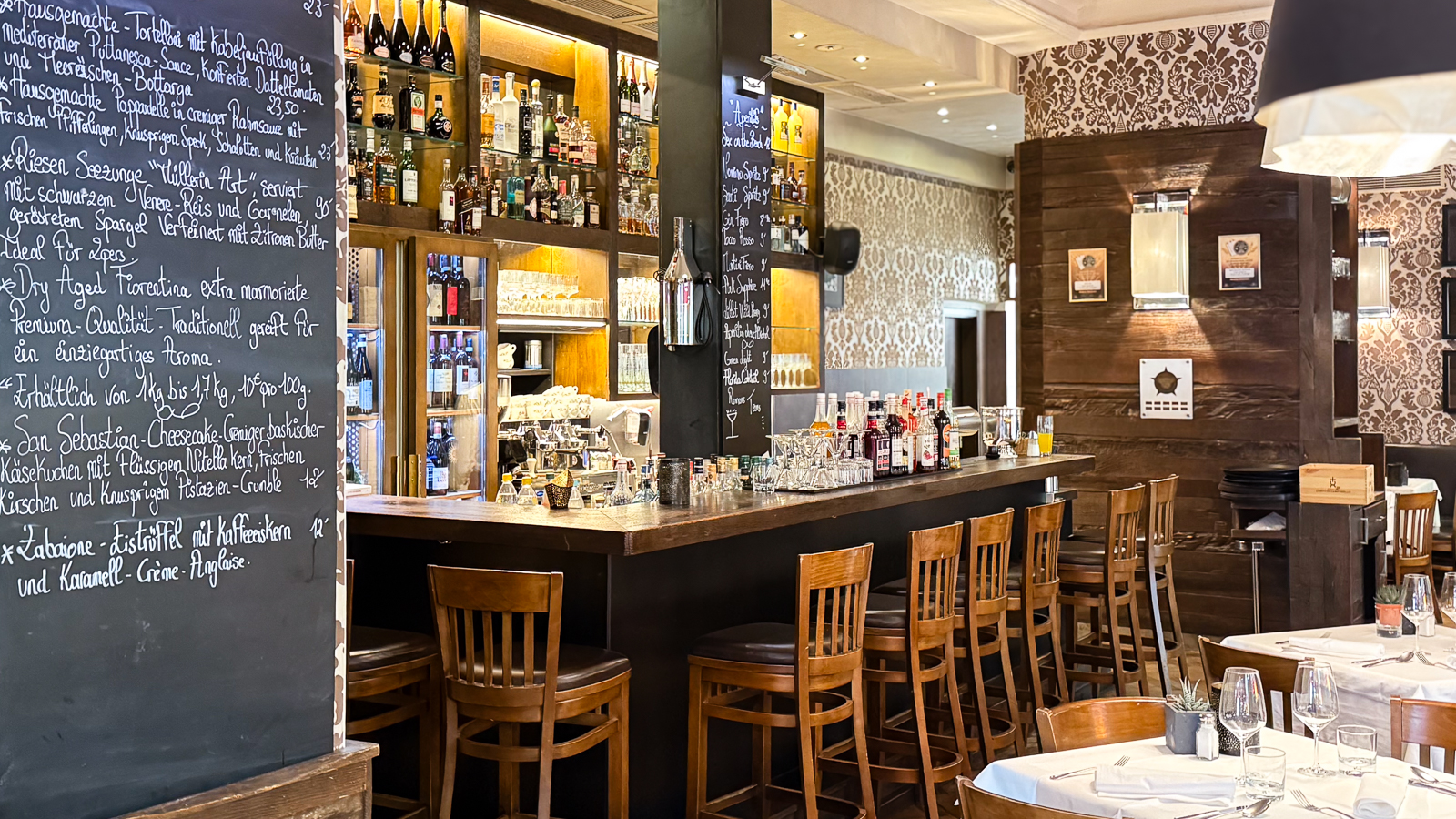 Der stilvoll eingerichtete Bar- und Restaurantbereich verbindet eine lange Holztheke mit beleuchteten Spirituosenregalen und gemütlichen Sitzplätzen. Gemusterte Tapeten, gedeckte Tische und warme Beleuchtung schaffen eine elegante und einladende Atmosphäre.