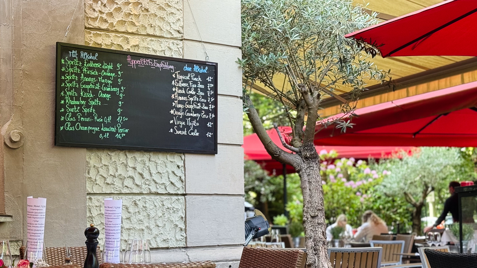 An einer hellen Hauswand hängt eine schwarze Kreidetafel mit den Aperitif-Empfehlungen des Restaurants. Daneben lädt die begrünte Terrasse mit roten Sonnenschirmen, gedeckten Tischen und mediterranen Pflanzen zum Verweilen ein.