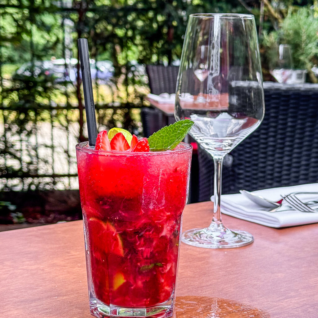 Auf einem gedeckten Terrassentisch steht ein leuchtend roter Caipiroska mit Erdbeeren in einem hohen Glas, garniert mit frischen Erdbeerscheiben und einem Minzzweig. Daneben stehen ein Weinglas sowie Besteck auf einer Serviette, während die grüne Restaurantterrasse im Hintergrund sanft unscharf erscheint.