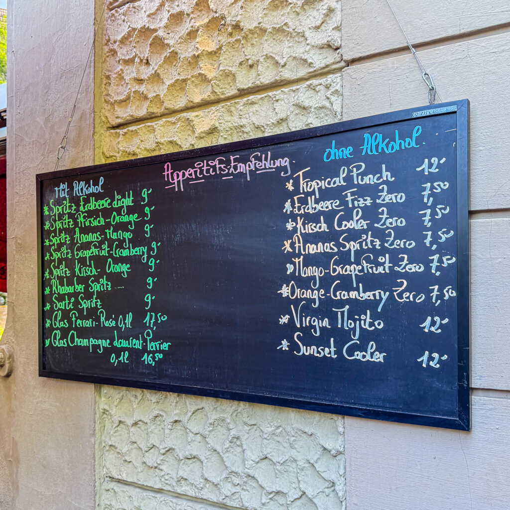 Eine schwarze Kreidetafel an einer hellen Hauswand zeigt die Aperitif-Empfehlungen des Restaurants. Die Getränkekarte ist in Kategorien für alkoholische und alkoholfreie Aperitifs unterteilt und mit farbiger Kreideschrift gestaltet.