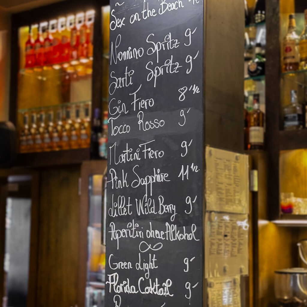 Eine schwarze Kreidetafel im Barbereich präsentiert eine Auswahl an Aperitifs und Cocktails in weisser Handschrift. Im Hintergrund sind beleuchtete Regale mit Spirituosen und Flaschen zu sehen, die eine gemütliche Bar-Atmosphäre schaffen.
