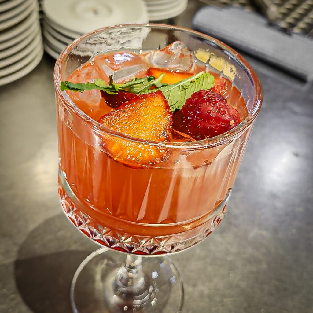 Ein ralkoholfreier Erdbeere Freeze Cocktail mit Eiswürfeln wird in einem eleganten Stielglas serviert und mit frischen Erdbeeren sowie einem Minzzweig garniert. Im Hintergrund sind gestapelte Teller und der Barbereich leicht unscharf zu erkennen.