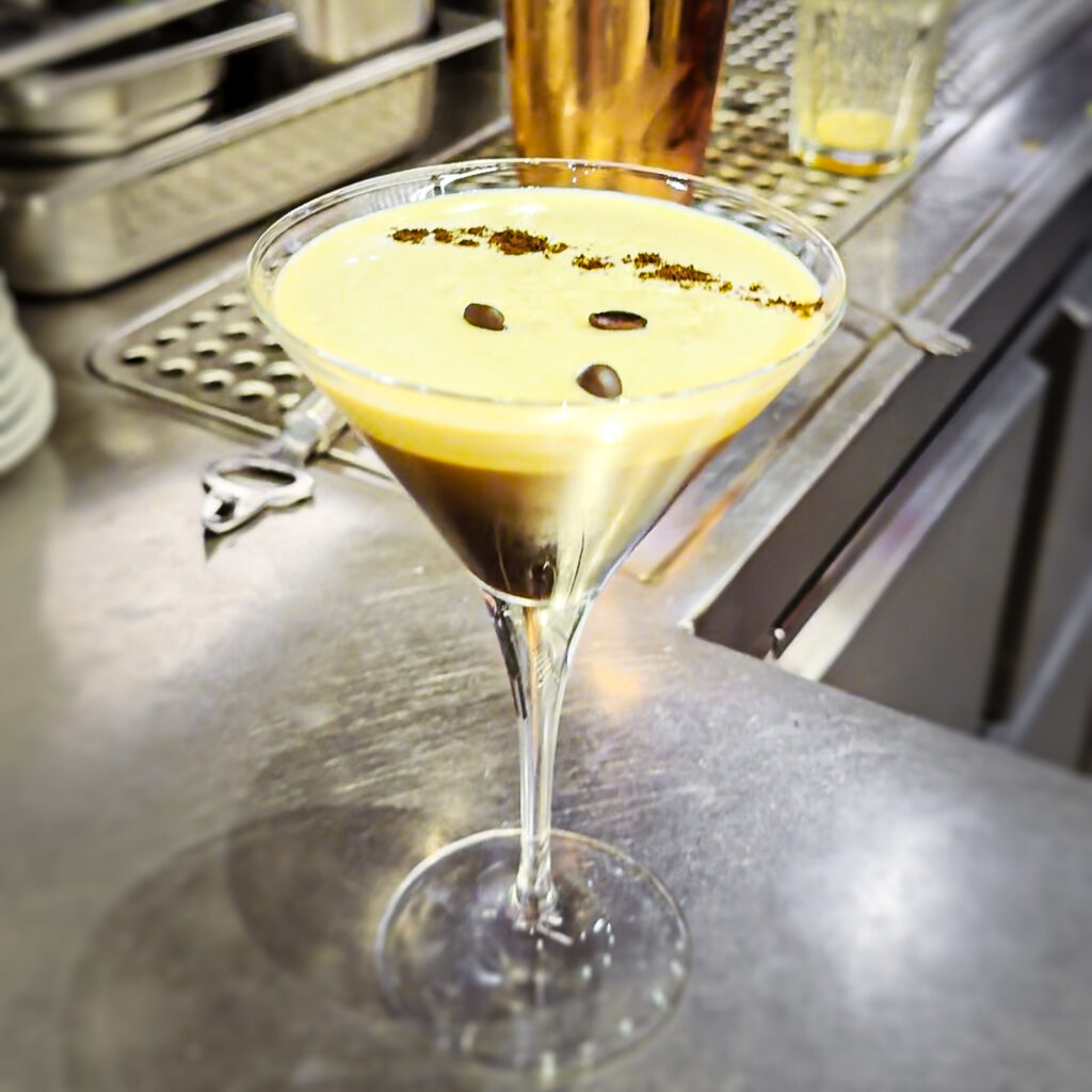 Ein cremiger Espresso Martini in einem klassischen Martiniglas steht auf einer Edelstahl-Bar. Die helle Schaumkrone ist mit Kaffeebohnen und feinem Kakaopulver dekoriert, während der Barbereich im Hintergrund leicht unscharf erscheint.