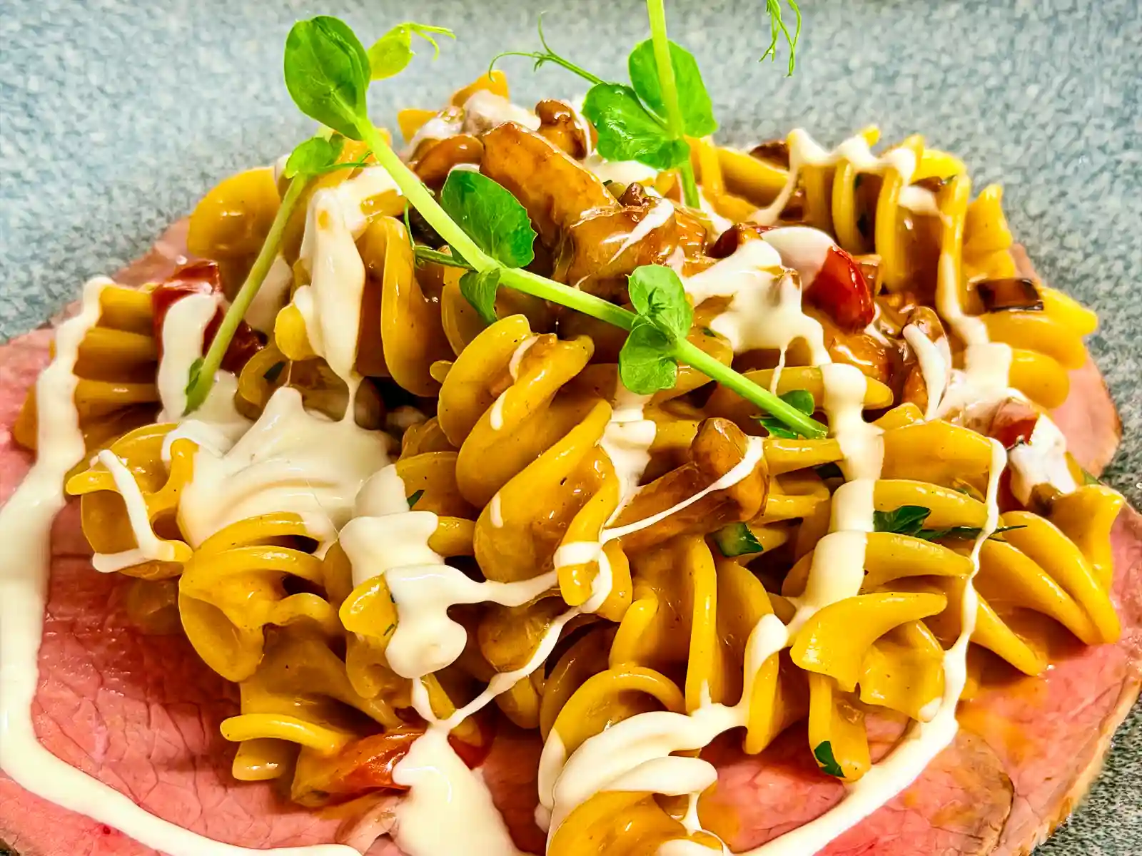 Pastagericht mit spiralförmigen Nudeln, brauner Sauce, weisser Creme, Erbsensprossen und rosa Fleischscheiben auf einem blau-grauen Teller.
