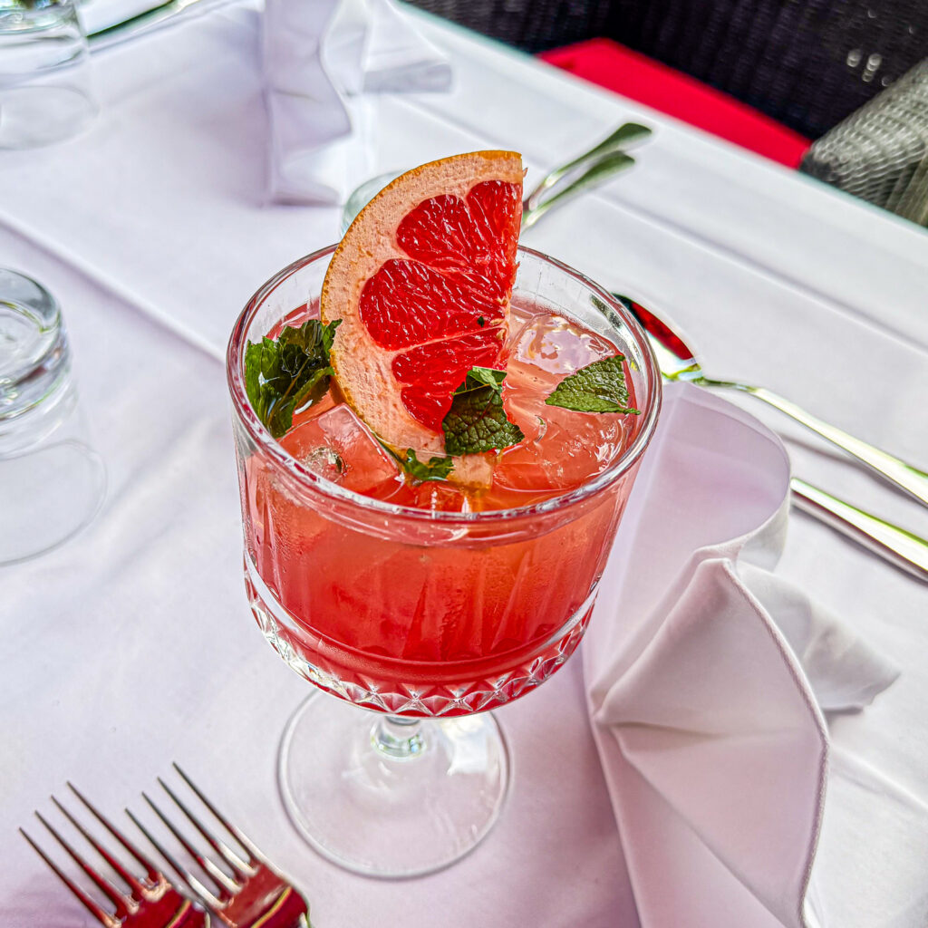 Ein Grapefruit Cranberry Cocktail mit Eiswürfeln wird in einem eleganten Stielglas serviert und mit einer grossen Grapefruitscheibe sowie frischer Minze garniert. Das Getränk steht auf einem weiss gedeckten Terrassentisch mit Besteck und gefalteten Stoffservietten.