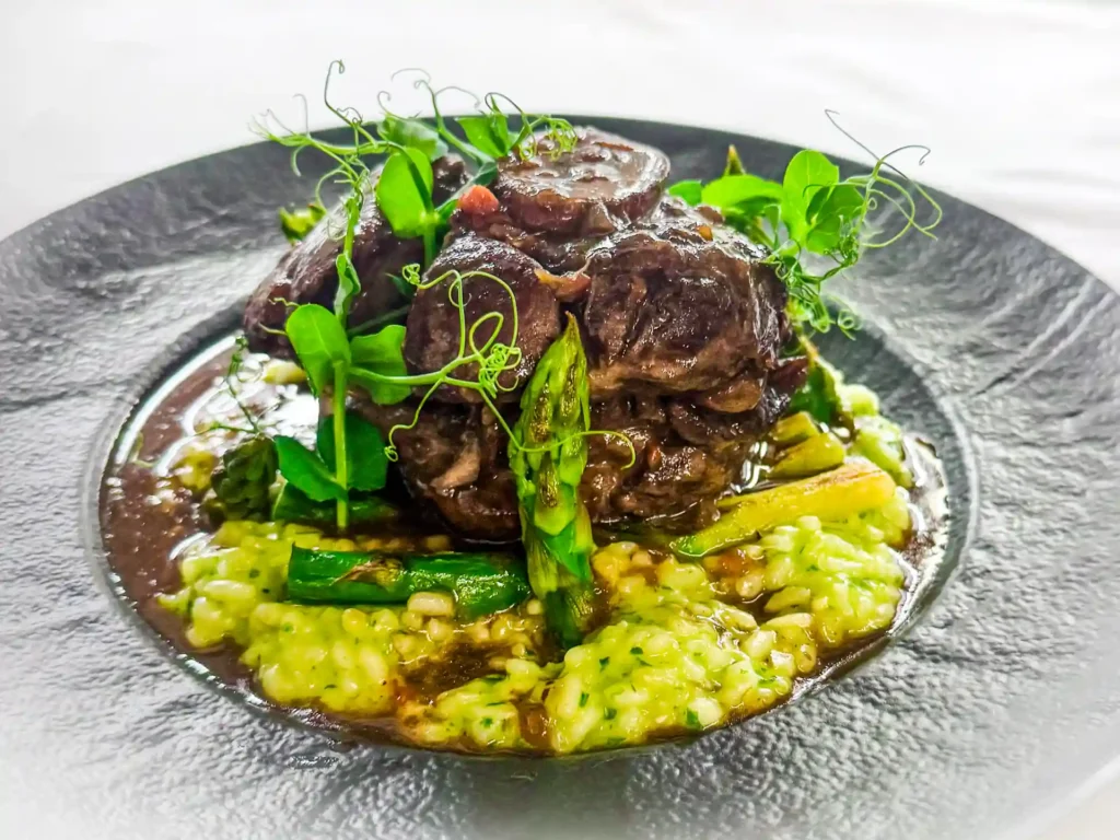 Ossobuco auf Risotto angerichtet auf einem schwarzen Teller.