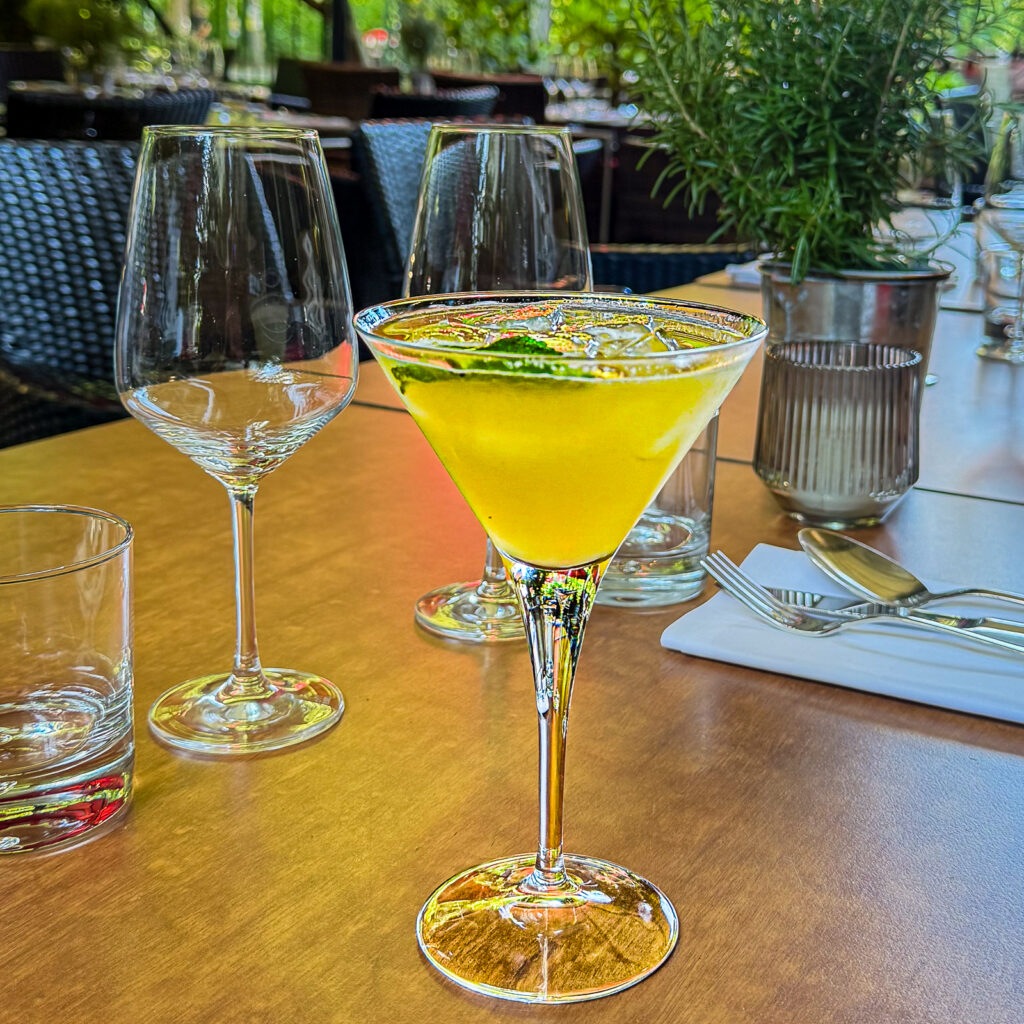 Ein Passion Martini Cocktail wird in einem eleganten Martiniglas serviert und mit einem Minzeblatt garniert. Das Getränk steht auf einem Holztisch auf der Restaurantterrasse, umgeben von Wein- und Wassergläsern, Besteck und einem Topf mit frischem Rosmarin.