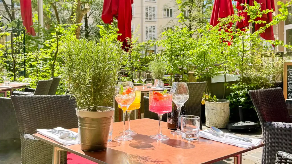 Auf einem sonnigen Terrassentisch stehen zwei farbenfrohe Cocktails in eleganten Gläsern, umgeben von Wein- und Wassergläsern sowie einer dunklen Flasche. Ein Topf mit frischem Rosmarin steht im Vordergrund, während die begrünte Restaurantterrasse mit roten Sonnenschirmen im Hintergrund eine einladende, mediterrane Atmosphäre schafft.