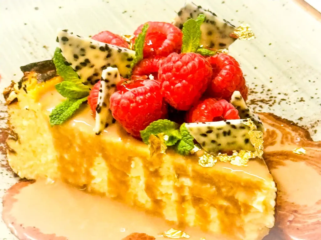 Stück San-Sebastian-Käsekuchen, garniert mit frischen Himbeeren, Drachenfruchtstücken, Minze und Goldblättchen, angerichtet mit Sauce auf einem Teller.