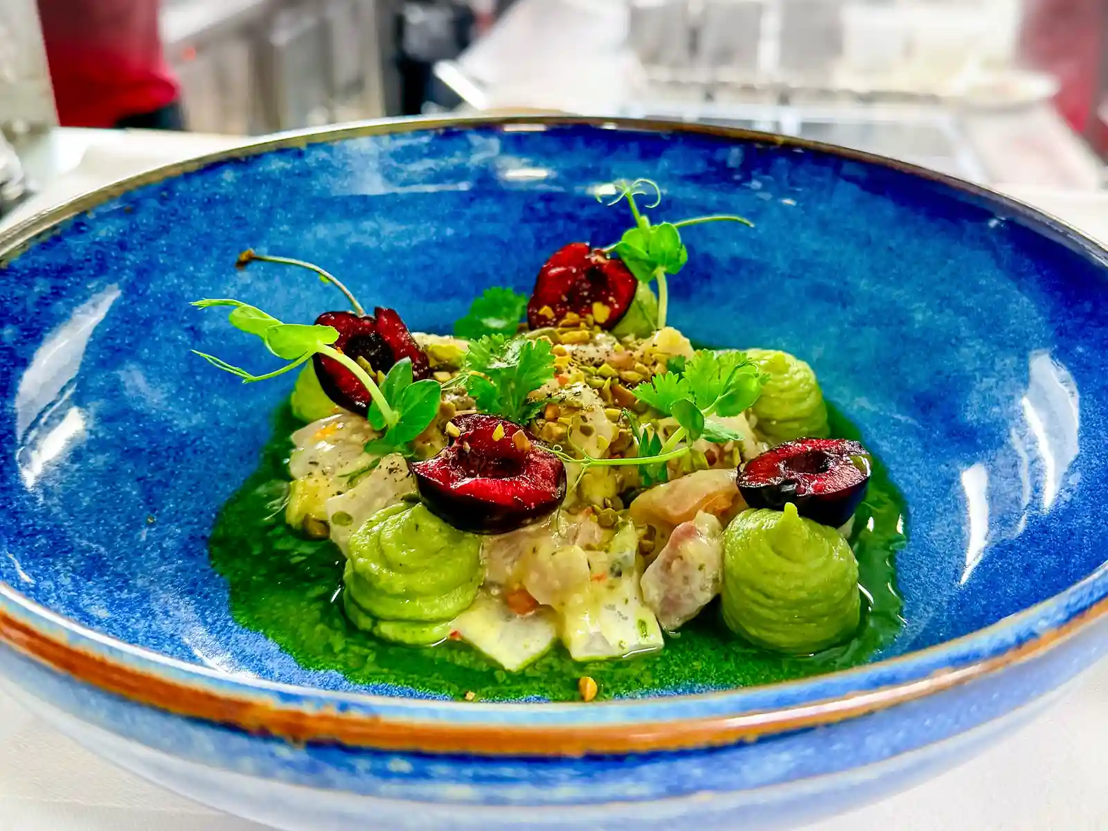 Device vom Wolfsbarsch in einer blauen Schale mit grüner Sauce, hellen Stücken, Avocado-Creme, halbierten Kirschen, Kräutern, Sprossen und gehackten Nüssen.