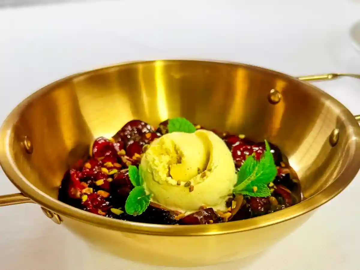 Flambierte Kirschen mit Quignolet und gerösteten Pistazien, angerichtet mit einer Kugel Vanilleeis und frischer Minze in einer Kupferpfanne.