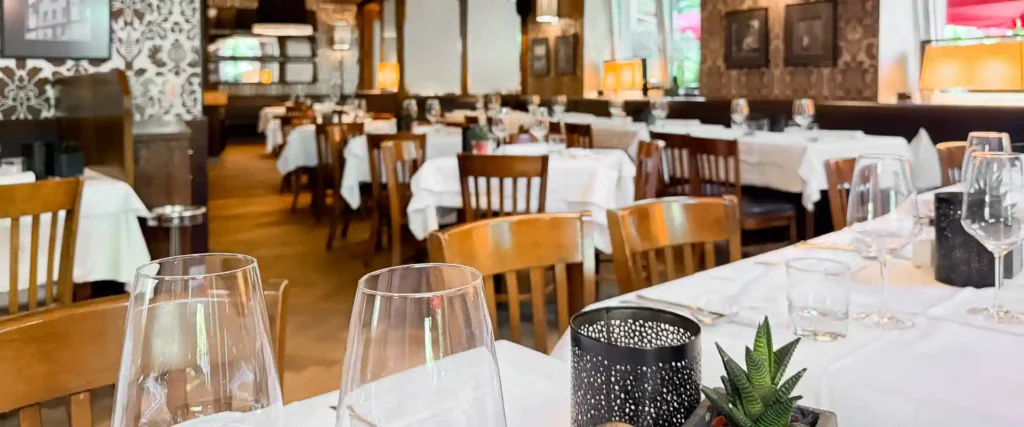 Restaurant-Innenraum mit gedeckten Tischen, Weingläsern, Holzstühlen, weissen Tischdecken und warmen Lampen.