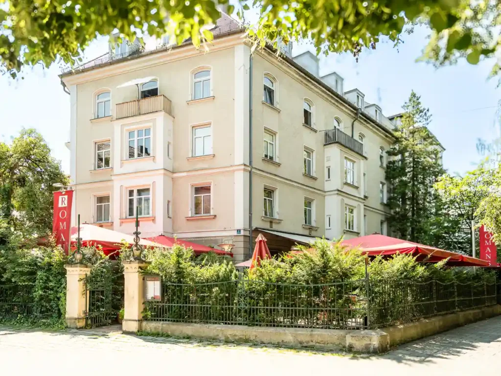 Helles mehrstöckiges Eckgebäude mit Ristorante Romans, rotem Sonnenschirmgarten und üppiger Begrünung an einem sonnigen Tag