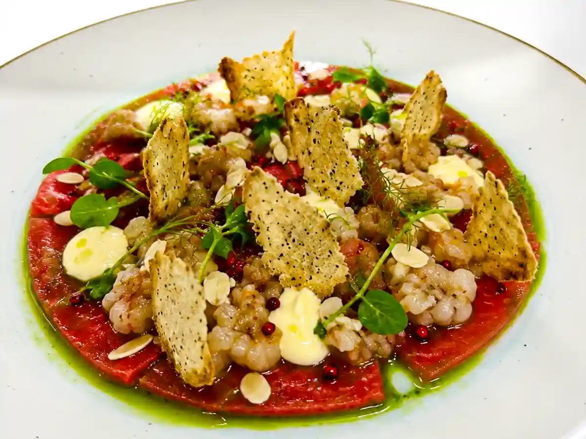Rohe Rotgarnele auf rund angerichtetem Wassermelonen-Carpaccio, garniert mit knusprigen Crostini, Yuzu-Mayonnaise, Datteltomaten-Gel, Basilikumöl, Limettenzeste, rosa Pfeffer, Mandelblaettchen und frischen Microkraeutern.