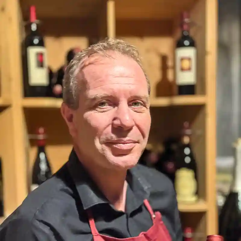 Porträt eines Sommeliers mit roter Schürze vor einem Holzregal mit Weinflaschen.