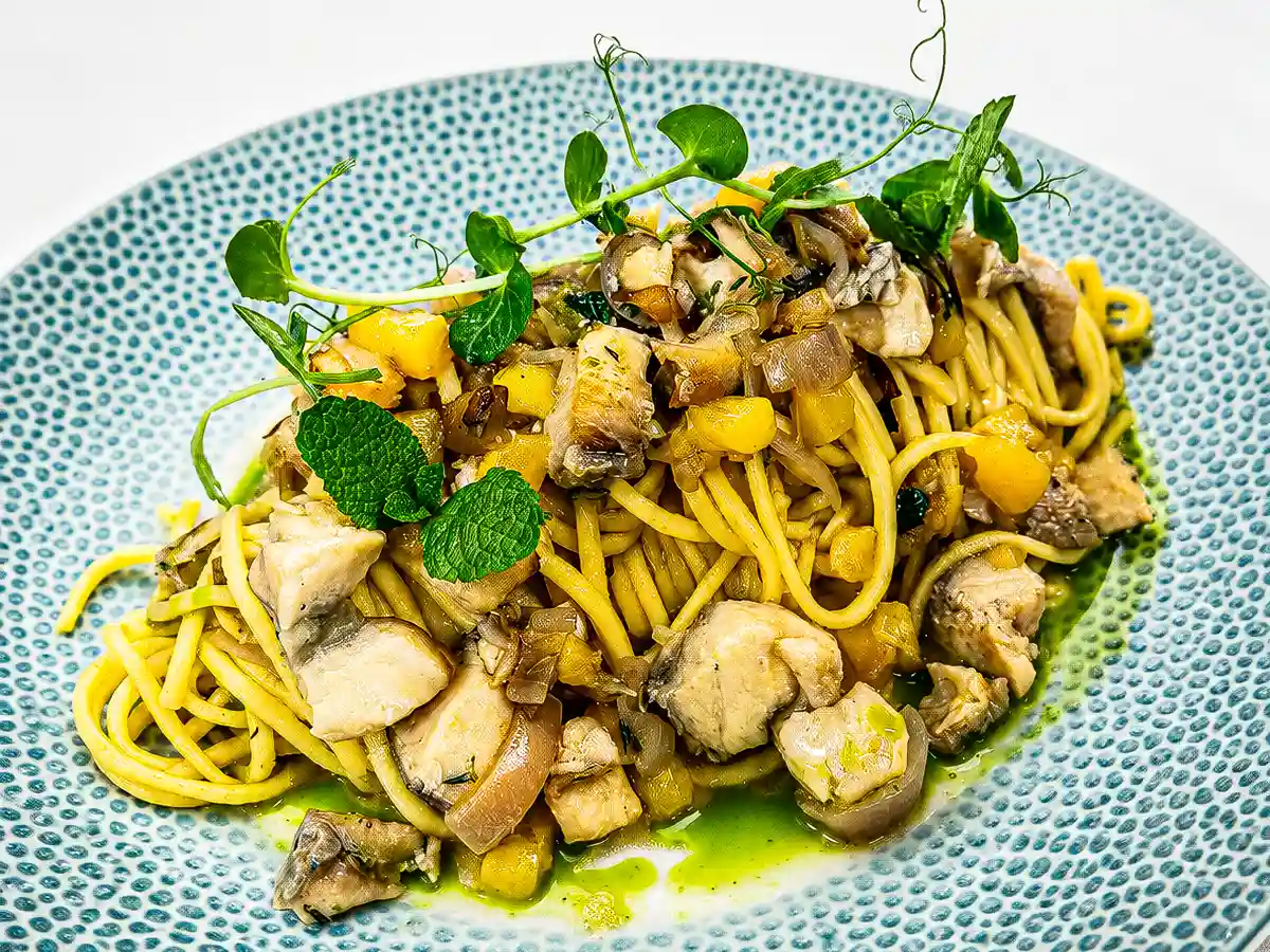 Cremig angerichtete Spaghetti mit Stuecken von Zackenbarsch und gelbem Pfirsich in einer feinen Kraeuterreduktion, garniert mit Minze, Microgreens, Limettenzeste und Olivenoel auf einem blau gemusterten Teller.