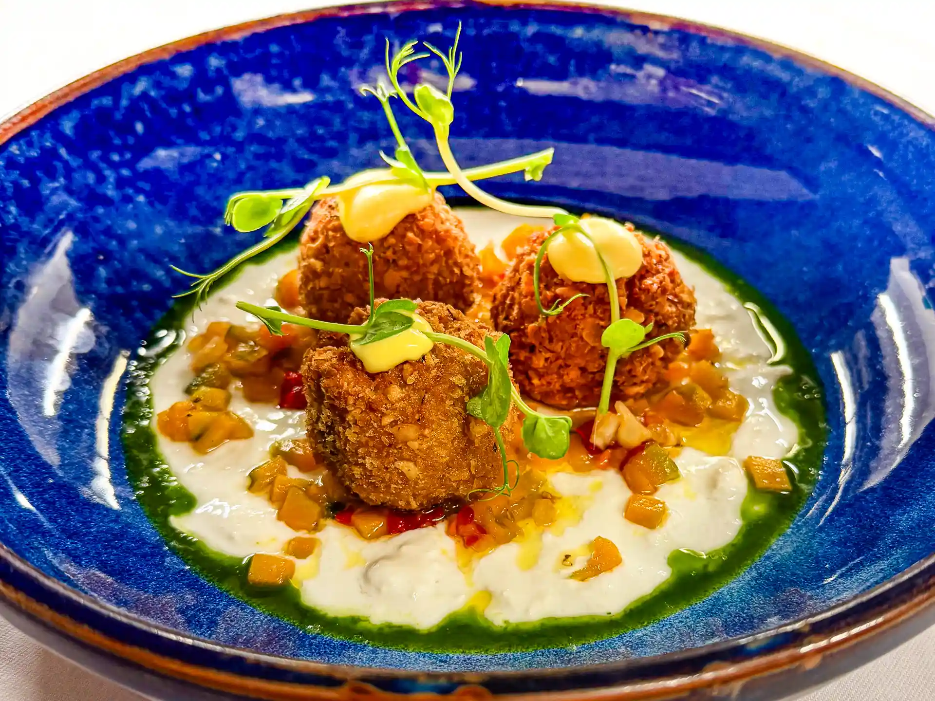 Vier knusprig panierte Bällchen auf cremiger Sauce mit Gemüsewürfeln, grünen Kräutersprossen und gelben Sauce-Tupfen in einem tiefblauen Teller.