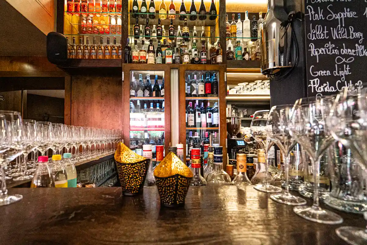 Gut ausgestattete Restaurantbar mit zahlreichen Flaschen in beleuchteten Regalen, Reihen sauber aufgestellter Weingl&auml;ser sowie einer beschrifteten Getr&auml;nketafel im Hintergrund.