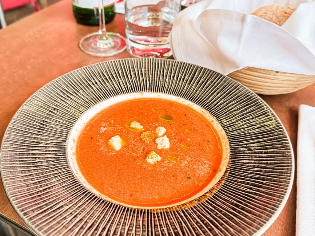 Nahaufnahme einer cremigen roten Suppe in einem grossen, geriffelten Teller, garniert mit kleinen Brotwürfeln und einigen Tropfen Öl; im Hintergrund stehen ein Brotkorb sowie Wasser- und Weingläser.
