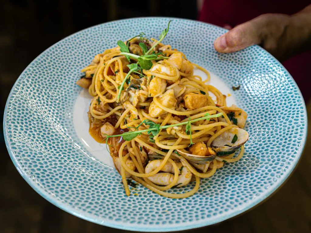 Nahaufnahme eines Pastagerichts mit Spaghetti, Muscheln und weiteren Meeresfrüchten in würziger Sauce, garniert mit frischen grünen Sprossen auf einem hellblauen Teller.
