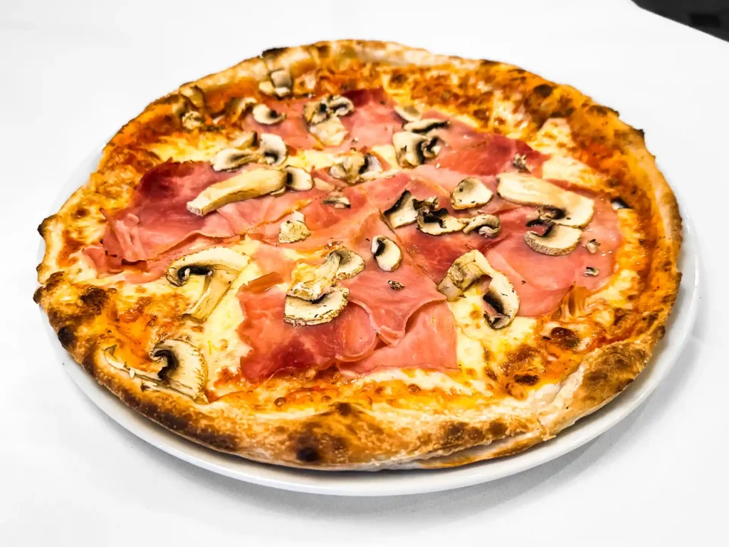 Pizza mit Schinken, Champignons, Tomatensauce und geschmolzenem Käse auf einem weissen Teller.