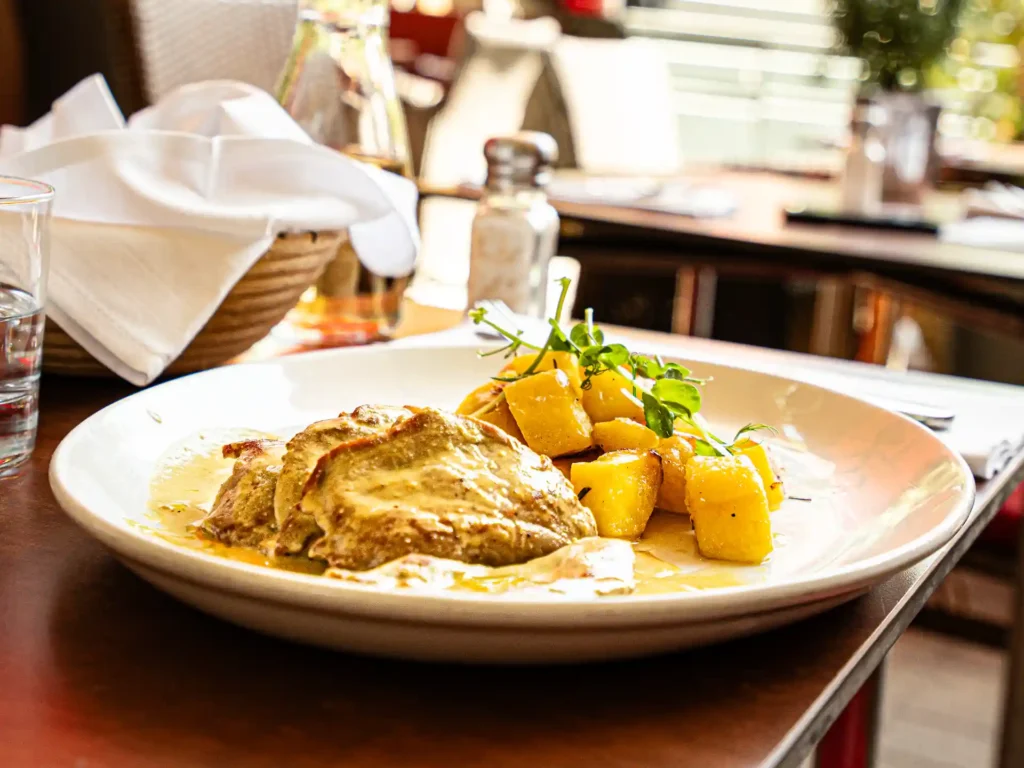 Nahaufnahme eines Restaurantgerichts mit goldbraun gebratenen Kartoffelstücken, frischen grünen Sprossen und Fleischstücken in heller Sauce; im Hintergrund stehen Wein- und Wassergläser auf dem Tisch.