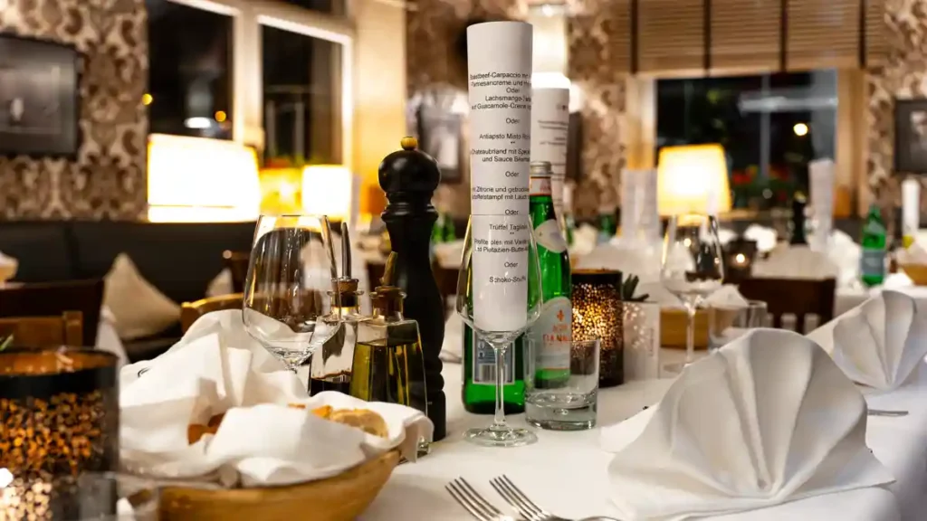 Festlich gedeckter Restauranttisch mit weisser Tischdecke, gefalteten Stoffservietten, Wein- und Wassergläsern, Brotkorb, Öl- und Essigflaschen sowie schwarzer Pfeffermühle; im Hintergrund ein warm beleuchteter Speiseraum.