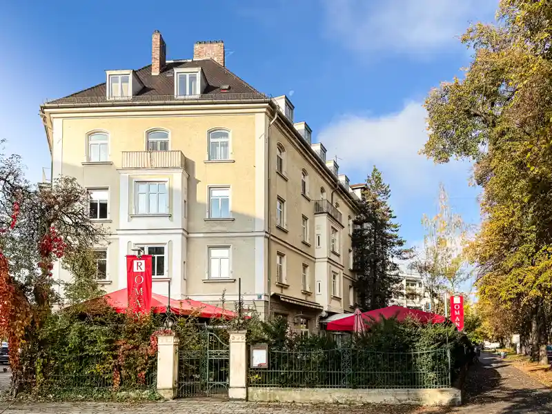 Aussenansicht des Ristorante ROMANS in München-Neuhausen. Das Restaurant befindet sich in einem mehrstöckigen, cremefarbenen Altbau mit roten Sonnenschirmen, begrüntem Aussenbereich und roten ROMANS-Schildern. Herbstlich gefärbte Bäume umrahmen das Gebäude bei blauem Himmel.